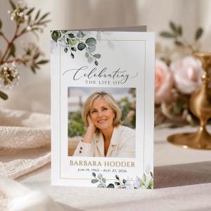 May include: A white funeral program card featuring a central photograph of a blonde woman, with the text "Celebrating THE LIFE OF", "BARBARA HODDER", and "JUN 19, 1962 - AUG 23, 2026" printed in black and gold, accented by green eucalyptus leaf illustrations.