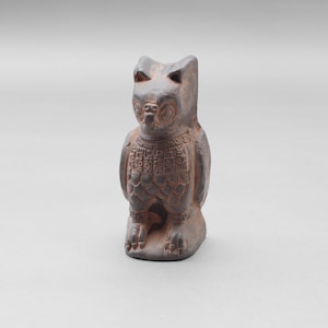 May include: A small, brown owl figurine with detailed carvings. The owl is depicted in a seated position with its wings folded. The figurine has a textured surface and is likely made of ceramic or a similar material. The owl's eyes and facial features are clearly defined.