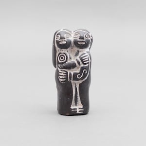 Puede incluir: Una pequeña escultura de color gris carbón que representa a dos figuras de pie, una al lado de la otra, en un abrazo. La pieza presenta líneas blancas incisas que definen los rasgos faciales, los brazos y los detalles de la ropa sobre la superficie mate.