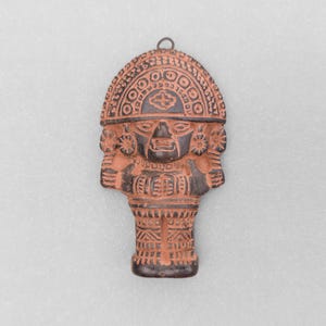 May include: A terracotta-coloured ceramic wall plaque featuring a stylised anthropomorphic figure with a large, semi-circular headdress. The figure displays intricate geometric carvings, dark brown accents, and a small metal loop at the top for hanging.