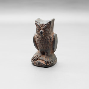 May include: A small, dark brown owl figurine with detailed carvings. The owl is perched on a small base, with its wings at its sides and its head upright. The figurine has a textured surface and is set against a plain background.