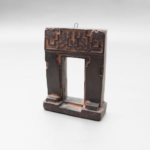May include: A dark brown, rectangular, decorative archway with an open centre. The top of the arch features carved geometric designs. The arch is made of a dark brown material with lighter brown accents, and has a small metal loop at the top.