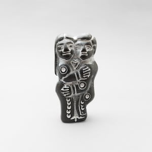 May include: A black carved sculpture of two embracing figures. The figures have stylized features and are joined together. A heart is carved between them. The sculpture is set against a white backdrop.