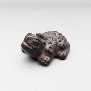 May include: A dark brown, carved wooden frog figurine. The frog has detailed features, including large eyes, a textured back, and visible legs. The figurine is set against a plain, light background.