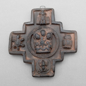 May include: A dark brown, cross-shaped decorative item with intricate carvings. The central design features a circular motif with figures, surrounded by other symbols including a turtle and an owl. A small loop is at the top for hanging.