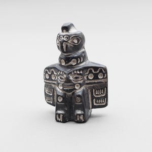 May include: A small, dark gray stone figurine carved in the shape of a bird with outstretched wings. The surface features intricate, light-colored etched patterns and lines that define the eyes, beak, and wing details on the front of the object.