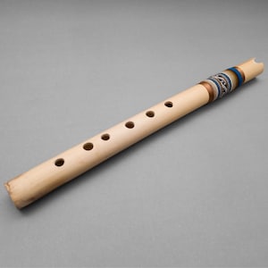 Op de afbeelding: Een houten fluit met zes vingergaten en een decoratieve band van blauw, wit en gouden garen. De fluit is gemaakt van natuurlijke bamboe.