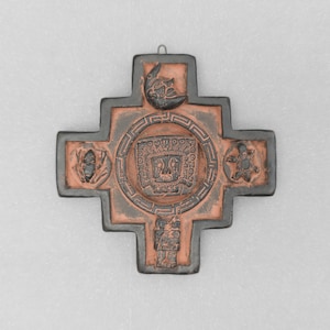 May include: A cross-shaped wall plaque featuring a dark brown and terracotta colour scheme. The centre displays a square face motif surrounded by a circular geometric border. Additional relief carvings include a crescent moon, a turtle, and figures.