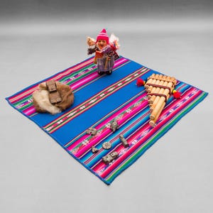 Peut inclure: Un tapis tissé coloré avec des rayures roses, bleues et blanches. Une petite poupée portant un chapeau rouge et une tenue traditionnelle péruvienne est placée sur le tapis. Une flûte en bois est également sur le tapis.
