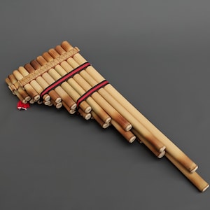 Op de afbeelding: Een traditionele Andes-panfluit gemaakt van bamboe met een rode en zwarte touwbinding. De panfluit ligt plat op een grijs oppervlak.
