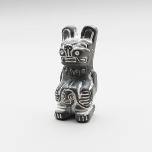 May include: A small, dark gray stone carving of a stylized bear figure with white etched details. The bear is depicted in a seated position, holding a smaller, circular face-like carving between its front paws, featuring distinct facial markings.