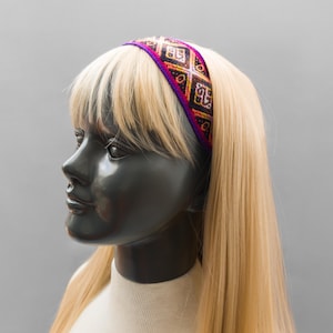 Pode incluir: Um busto de manequim preto com cabelo loiro comprido e uma faixa colorida. A faixa apresenta um padrão geométrico em roxo, dourado, vermelho e preto. O manequim está usando uma gola alta branca.
