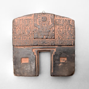 May include: A terracotta-coloured wall hanging with a central doorway and detailed relief carvings. The upper section features a central figure with a sun-like halo, flanked by detailed designs. The lower section is dark brown.