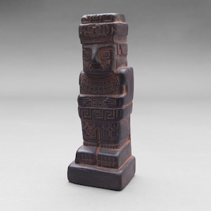 Könnte beinhalten: Eine dunkelbraune Steinskulptur einer stehenden Figur mit Kopfbedeckung und aufwendigen Schnitzereien. Die Figur hat einen quadratischen Sockel und ist mit geometrischen Mustern verziert.