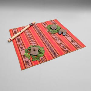 Peut inclure: Un tissu rectangulaire rouge à rayures verticales orange et rose avec des motifs marron foncé répétitifs, une flûte en bois clair avec des bandes sombres, deux tas de feuilles vertes et deux petits objets rectangulaires sculptés marron.