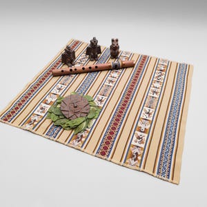Peut inclure: Un textile tissé rectangulaire avec des rayures verticales beiges, bleues et rouges ornées de motifs d'oiseaux et géométriques. Une flûte en bois, trois petites figurines d'animaux sculptées et une plaque solaire sur des feuilles vertes.