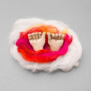 May include: A small sculpture of two hands with fingers extended, set within a vibrant arrangement of white, pink, orange, and yellow fibers. The hands are a light beige color, and the fibers create a colorful, textured background.