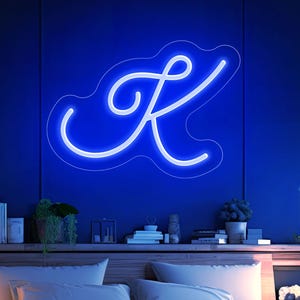 Puede incluir: Un letrero de neón azul con la forma de la letra "K" está montado en una pared azul oscuro sobre una cama. El dormitorio tiene una cabecera de madera, mesitas de noche y una estantería. La cama está hecha con almohadas blancas y una manta azul.