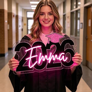 May include: A graduation sign with the name "Emma" and the year "2026" in a neon pink glow. The sign features a graduation cap with pink glitter. The sign is held in front of a person wearing a graduation gown.