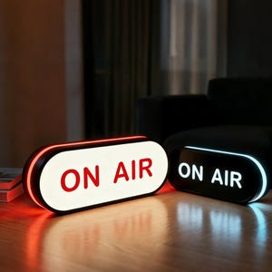 May include: Two illuminated "ON AIR" signs. One is white with red text and a red border, the other is black with white text and a blue border. Both are rectangular with rounded corners, resting on a wooden surface.