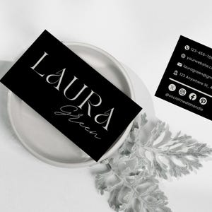 Op de afbeelding: Zwarte visitekaartjes met witte tekst, met de naam "LAURA Green" in elegante lettertypen. E&eacute;n kaart ligt op een wit bord, terwijl een andere contactgegevens weergeeft, waaronder een telefoonnummer, website en social media handle.