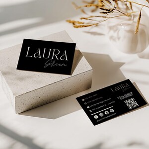 Op de afbeelding: Twee zwarte visitekaartjes met witte tekst en een QR-code. De kaarten tonen de naam "LAURA Green" in elegant schrift. E&eacute;n kaart rust op een wit, getextureerd blok, terwijl de andere plat ligt. Contactgegevens en social media-handles zijn ook zichtbaar.