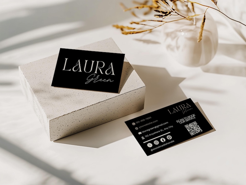 Op de afbeelding: Twee zwarte visitekaartjes met witte tekst en een QR-code. De kaarten tonen de naam "LAURA Green" in elegant schrift. E&eacute;n kaart rust op een wit, getextureerd blok, terwijl de andere plat ligt. Contactgegevens en social media-handles zijn ook zichtbaar.