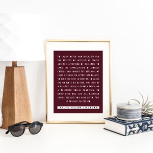May include: A framed print with a burgundy background and white text. The quote reads: "To laugh often and much, to win the respect of intelligent people and the affection of children, to earn the appreciation of honest critics and endure the betrayal of false friends; to appreciate beauty, to find the best in others, to leave the world a bit better, whether by a healthy child, a garden patch, or a redeemed social condition; to know even one life has breathed easier because you have lived, this is to have succeeded." The quote is attributed to Ralph Waldo Emerson.