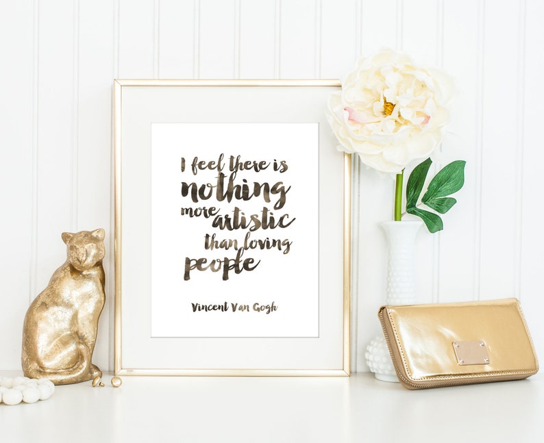 May include: A gold framed print with a white background and a quote in black script that reads "I feel there is nothing more artistic than loving people - Vincent Van Gogh". The print is sitting on a white surface with a gold cat figurine, a white vase with a white flower, and a gold clutch.