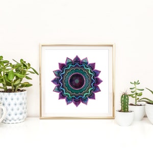 May include: A framed print of a colorful mandala with a purple, green, and blue color scheme. The mandala is surrounded by a white background and a gold frame. The print is displayed on a shelf with several potted plants.