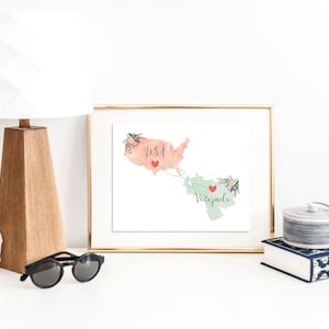 May include: A framed print with a watercolor map of the USA and Venezuela connected by a dotted line with hearts. The USA is in peach and Venezuela is in green. The text "USA" and "Venezuela" are written on the map.