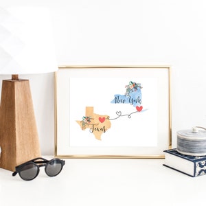 K&ouml;nnte beinhalten: Ein goldfarbener Druck mit einer Aquarell-Illustration der Bundesstaaten Texas und New York, die durch eine Linie mit Herzen verbunden sind. Der Text "Texas" und "New York" steht auf den Bundesstaaten.