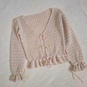 Könnte beinhalten: Cremefarbene gehäkelte Strickjacke mit langen Ärmeln und V-Ausschnitt. Die Strickjacke hat einen Bindverschluss vorne und Rüschendetails an Saum und Manschetten. Das offene Webmuster ist sichtbar.