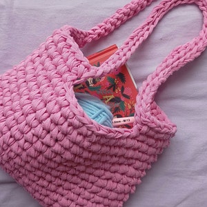 Op de afbeelding: Roze gebreide tote bag met een boek en garen erin. De tas heeft een lange band en is gemaakt van dik garen.