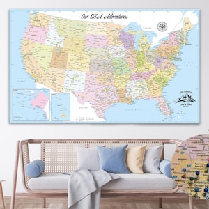 May include: A large, rectangular map of the USA with state borders in various colors. The map is labeled "Our USA Adventures" and includes a compass rose and mountain graphic. The map is displayed above a sofa with pillows.