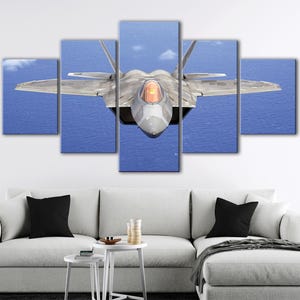 May include: A five-panel canvas print of a gray fighter jet in flight over a blue ocean. The jet's cockpit is orange, and the wings are angled. The artwork is displayed above a white couch with black pillows.