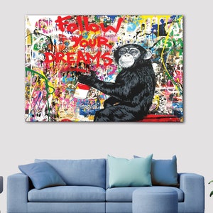 May include: A colourful abstract painting featuring a chimpanzee holding a red paint can. The chimpanzee is sitting on a red platform. The painting includes the words "Follow Your Dreams" in red paint.