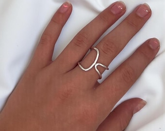 May include: A silver-colored ring with a unique open design, featuring two curved bands that intersect. The ring is worn on a hand with manicured nails, set against a white fabric background.