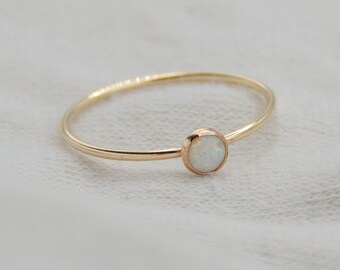 May include: A delicate gold ring with a small, round opal gemstone. The ring has a thin band and a simple, elegant design. The opal has a milky white color and is set in a bezel setting.