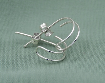 May include: A pair of small, silver hoop earrings. The earrings have a simple, minimalist design with a curved, double-hoop shape. The post and backing are also visible. The earrings are displayed on a light green fabric background.