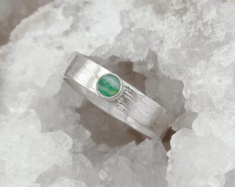 May include: A silver ring with a textured band and a round, green gemstone. The ring is set against a backdrop of white, crystalline material, highlighting its design and color.