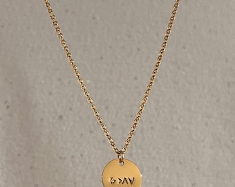 May include: Gold-colored necklace with a delicate chain and a round pendant. The pendant features the engraved text "AV>4". The necklace is displayed against a neutral background, highlighting the jewelry's design.
