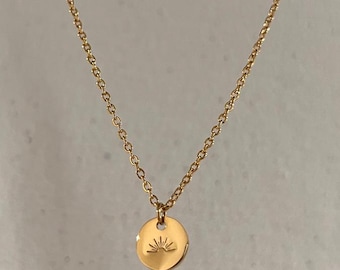 May include: A gold-colored necklace featuring a delicate chain and a circular pendant. The pendant has a sunrise design etched into its surface. The necklace is displayed against a neutral background, highlighting its simple elegance.