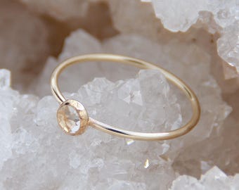 May include: A delicate gold ring featuring a round, light-colored gemstone. The ring's band is thin and polished, and the gemstone is set in a simple bezel. The ring is displayed on a white, crystalline surface, creating a contrast between the warm gold and the cool background.