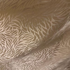 May include: Close-up of a beige fabric with a repeating pattern of stylized leaf shapes. The fabric is slightly translucent, with the pattern appearing in a lighter tone against the base color. The fabric is draped, creating shadows and highlights.