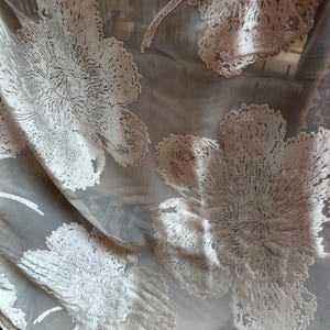 May include: A sheer, translucent fabric featuring large, raised white floral embroidery with intricate, textured petal details. The delicate mesh base provides a light, airy quality, while the dense, stitched floral patterns create a prominent, tactile contrast against the semi-transparent background material.