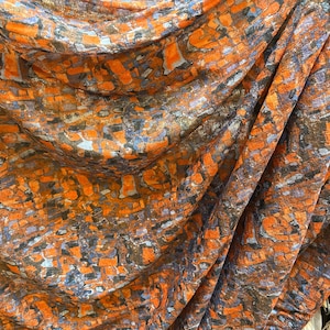 May include: Close-up of a draped fabric with an abstract pattern. The design features a mosaic of orange, brown, gray, and blue shapes. The fabric has a soft, flowing texture, with folds and shadows adding depth to the design.