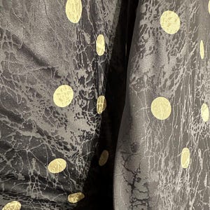May include: Close-up of a black fabric with a textured, crackled pattern and large, gold-colored polka dots. The fabric is folded, revealing the interior. The overall effect is one of luxury and sophistication.