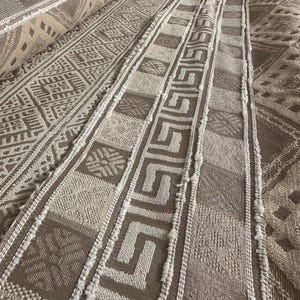 May include: Close-up of a beige and brown woven fabric with a geometric pattern. The fabric has a textured surface and a fringe edge.