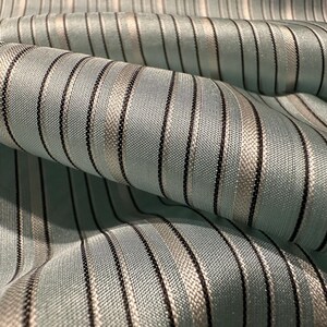May include: Close-up of a folded fabric with vertical stripes. The fabric has a light blue background with alternating black and silver stripes. The fabric appears to be woven with a subtle sheen, suggesting a luxurious texture.